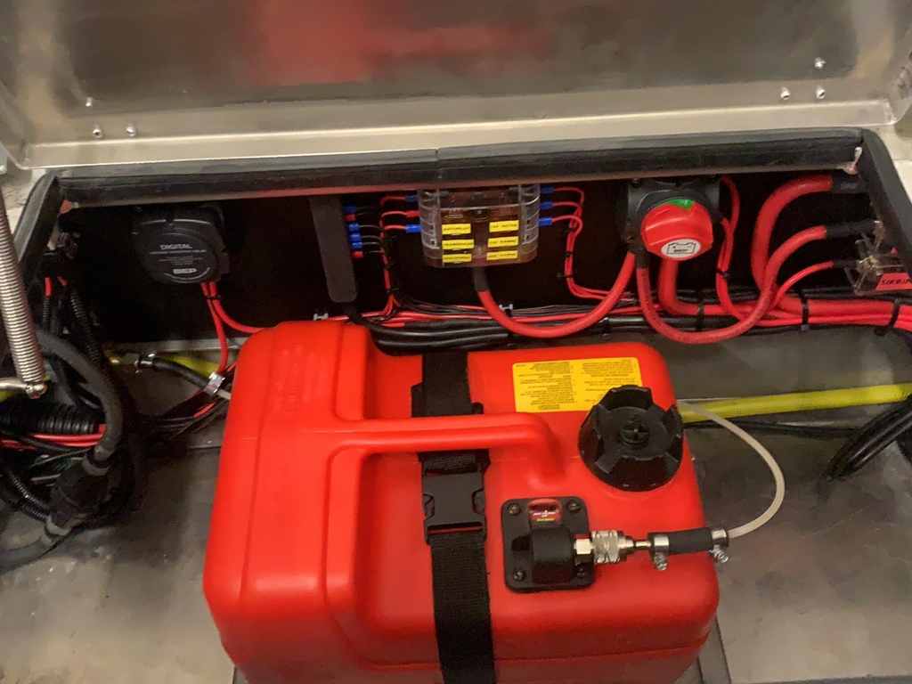 Åbent rum i en båd med en rød brændstoftank spændt fast, en mærket sikringsboks med blå og gule klemmer, røde batterikabler og en hovedafbryder